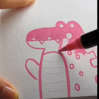 A close-up of a hand using a dual tip acrylic paint pen to add colorful details to a pink dinosaur drawing on paper.