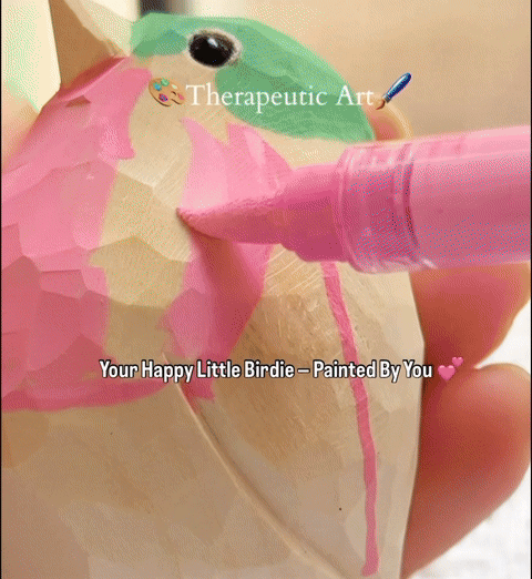 A person's hands using blue and pink acrylic paint markers to color a natural unfinished wooden bird, part of a DIY wooden animal coloring kit.