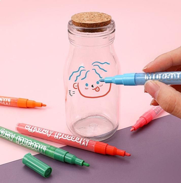 A person using a blue hard-tip acrylic marker to draw on a glass jar with colorful markers surrounding it.