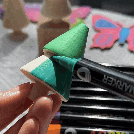 A person coloring a wooden Christmas tree with green paint using a marker, surrounded by colorful craft materials.