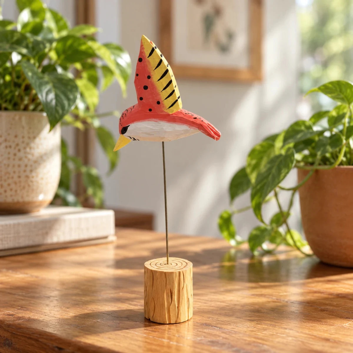 Red and yellow hand-painted wooden hummingbird ornament on a wire stand beside houseplants