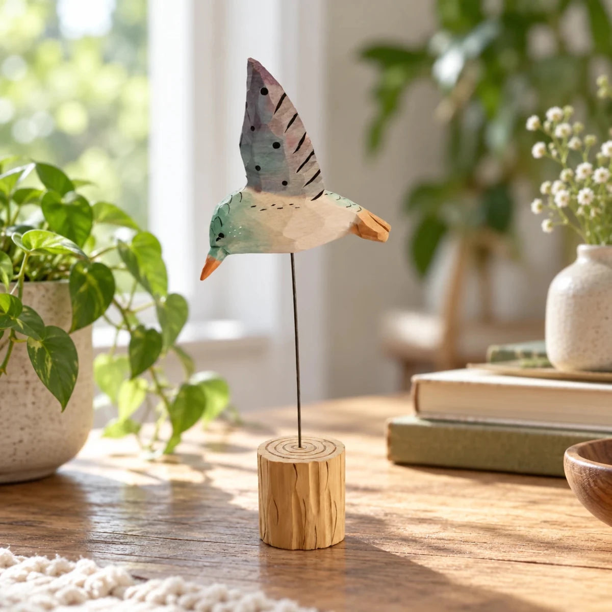 Sage green and white wooden hummingbird ornament on a wire stand beside plants and books